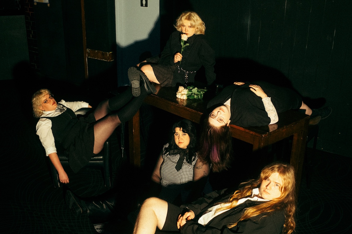 Promo photo of indie punk band Worm Girlz posing dramatically around a wooden table in dark, gothic-inspired outfits with pale roses.